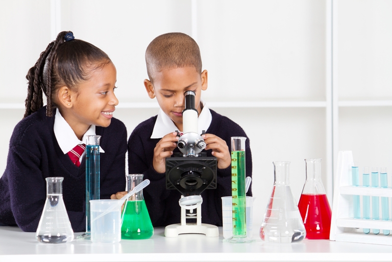 Two young students look at a microscope surrounded by beakers and test tubes. 